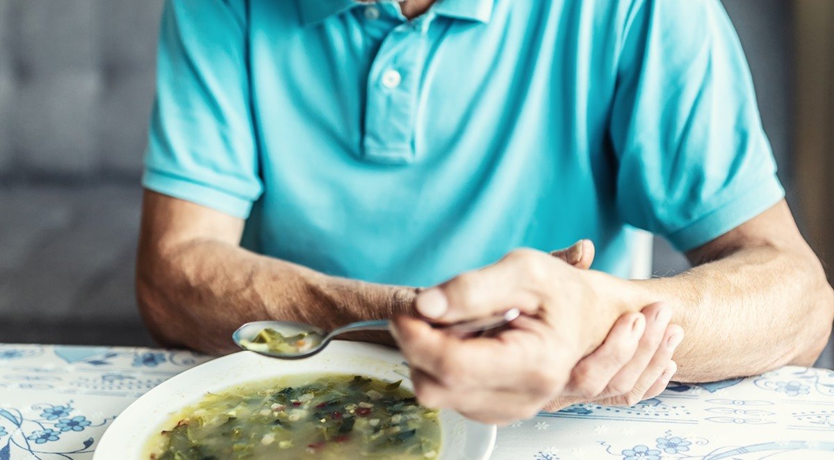 home idoso segura seu próprio braço com a outra mão para tentar comer uma sopa. ele tem Parkinson - Metrópoles