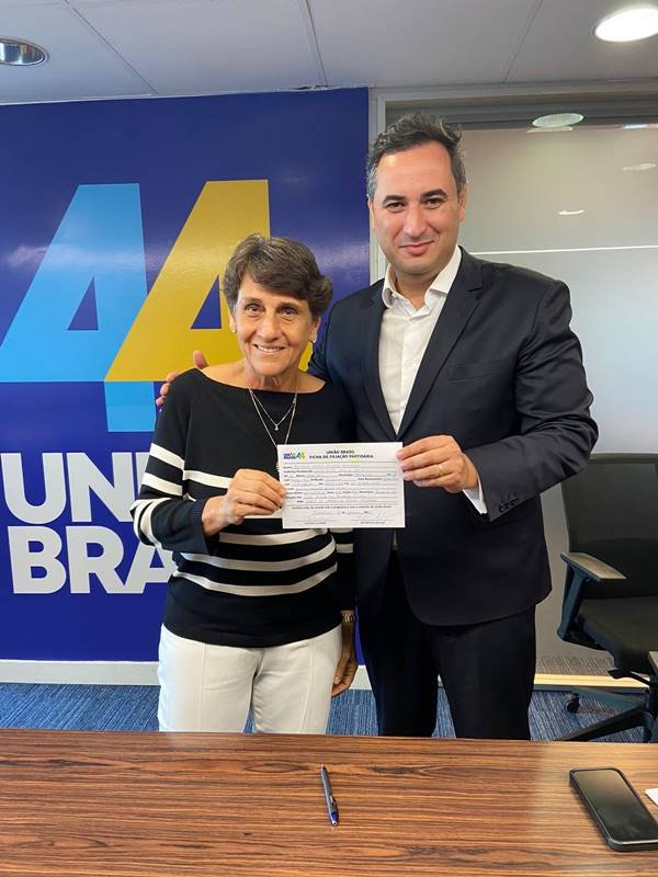 A política Eliana Pedrosa segura sua ficha de filiação ao União Brasil ao lado do presidente da legenda do DF. Ao fundo, um banner com o número da sigla - Metrópoles