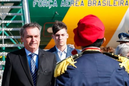 Bolsonaro e seu filho, Jair Renan, diante de militar em cerimônia após desembarcarem de avião da FAB. O vento bate no rosto deles e seu cabelo está eriçado - Metrópoles
