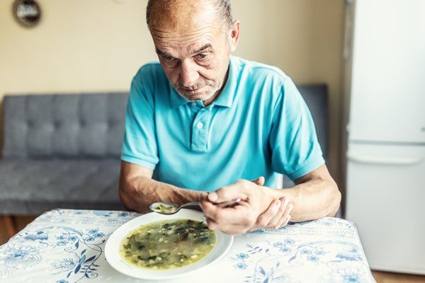 homem branco e idoso come uma sopa enquanto segura a própria mão. ele usa uma camisa polo azul