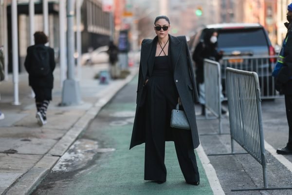 Em Nova York, mulher usa look todo preto, com calça de alfaiataria, blusa com alças finais transpassadas na frente e sobretudo