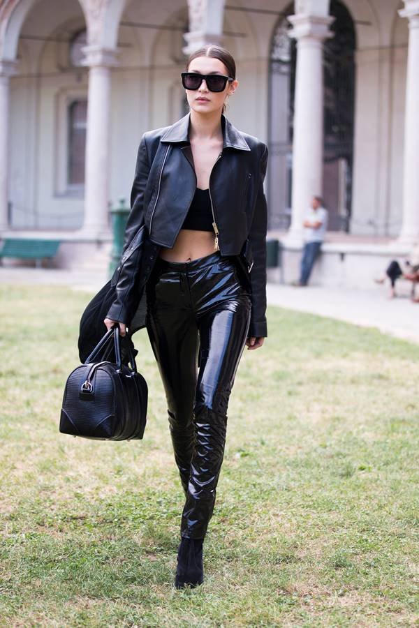Modelo Bella Hadid andando na grama. Ela usa look todo preto, com top, jaqueta e calça de couro, além de bolsa segurada na mão