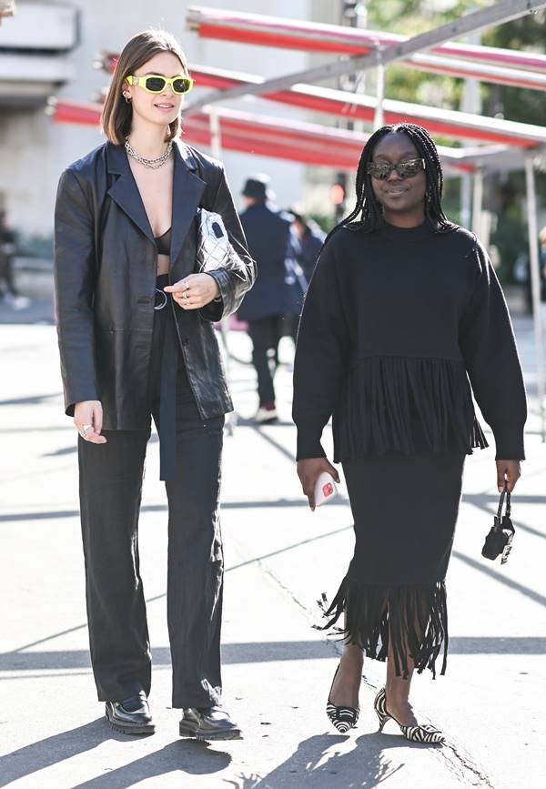 Duas mulheres caminham lado a lado. Uma é branca e usa top preto, com jaqueta preta de couro e top preto. A outra é negra e usa saia e casaco de moletom. Ambos com franjas na frente