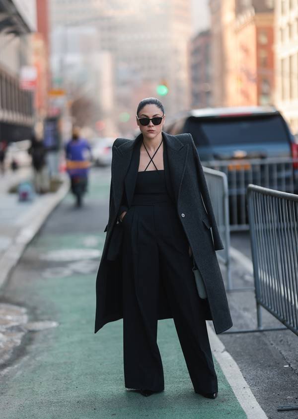 Em Nova York, mulher branca usa look todo preto, com calça de alfaiataria, blusa com alças finais transpassadas na frente e sobretudo