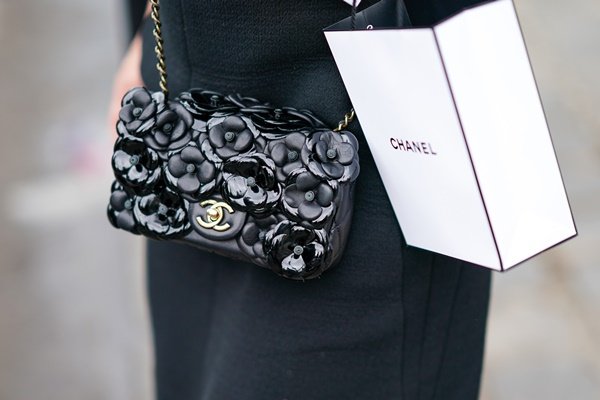 Bolsa preta da marca Chanel que imita a textura de várias flores Camélia. A pessoa segura uma sacola da mesma marca, a Chanel, como se tivesse saindo da loja 