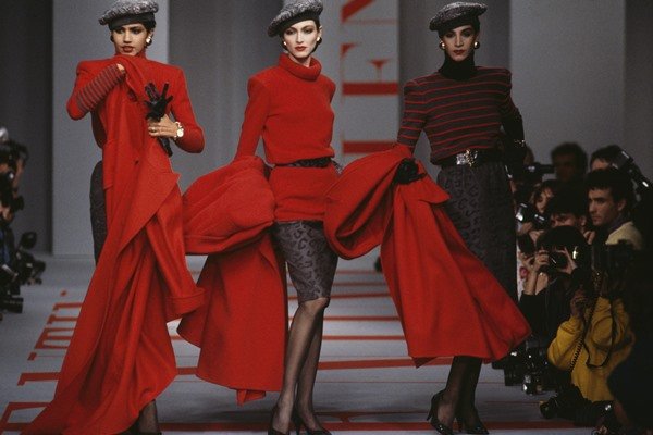 Desfile de 1986 da marca Valentino. Na passarela, três mulheres posam para a foto. As duas primeiras usam roupas vermelhas e a última usa um look preto, porém segura nas mãos um casaco também vermelho