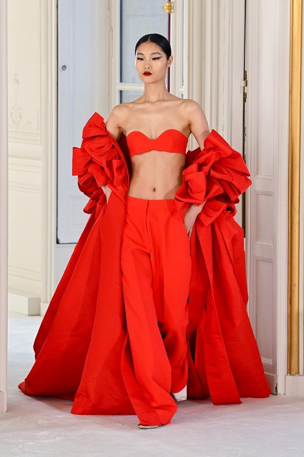 Mulher branca e asiática de cabelo preto preso em coque baixo. Ela desfila em coleção da Valentino e usa calça, top e casaco vermelho. 