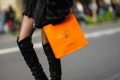 Mulher andando com sacola laranja da marca Hermès. Ela usa meia calça preta fina, uma bota preta de cano alto e um saia também preta.