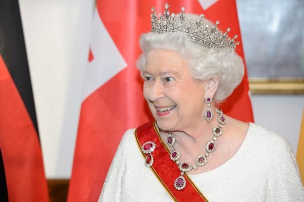 Foto colorida. Rainha Elizabeth com joias e uma faixa vermelha