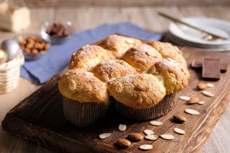Colomba Pascal caseira ao lado de amêndoas e chocolate sob tábua de madeira em mesa - Metrópoles