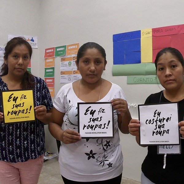 Três mulheres morenas segurando placas com os dizeres "Eu fiz a sua roupa". Elas possuem o cabelo liso amarrado e usam camisetas preto e brancas e calças jeans - Metrópoles 