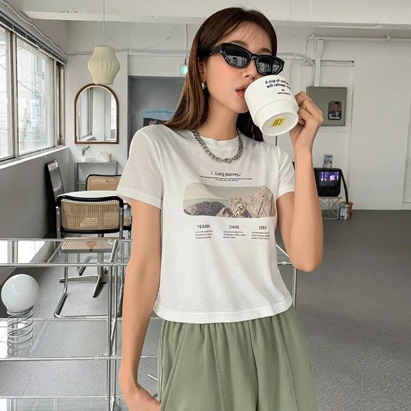 Mulher branca asiática com cabelo liso e longo. Ela está bebendo algo em uma caneca e usa óculos escuros, camiseta branca e uma calça de moletom verde. Todas as peças são da Shein.