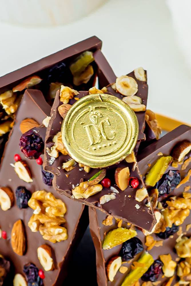 Barras de chocolate Dolce Cacao com castanhas e logo dourado da marca, também em chocolate - Metrópoles