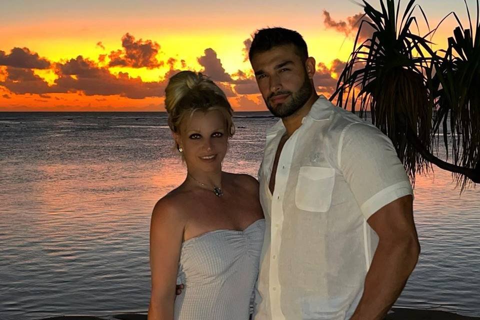 Foto colorida de Britney Spears e Sam Asghari em praia usando roupa branca - Metrópoles