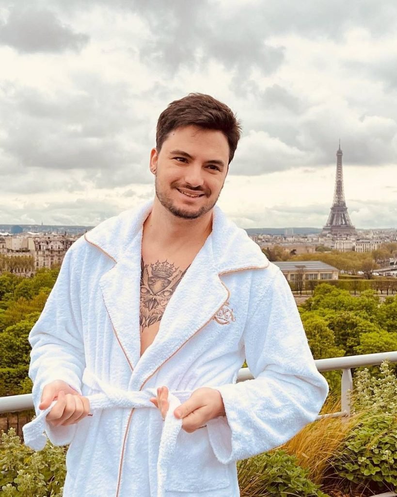 O youtuber Felipe Neto posa para foto e sorri usando um roupão branco na sacada de hotel de luxo em Paris. Ao fundo, é possível ver a torre Eiffel - Metrópoles