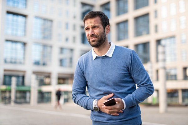 Man standing with one hand on his stomach and the other holding a cell phone.  He wears a blue T-shirt and has dark hair and beard - Metropolises