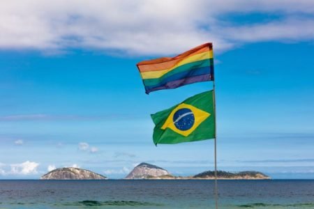 Bandeira LGBTQIA+ e bandeira do Brasil afixadas em mastro tremulam em praia. Ao longe, é possível ver mar e ilhas - Metrópoles