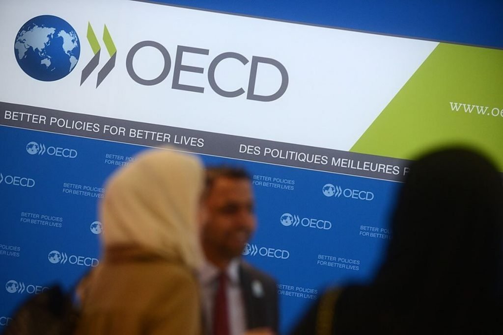 Imagem de reunião da OCDE, com pessoas conversando. Ao fundo, em um painel, o logotipo da entidade - Metrópoles