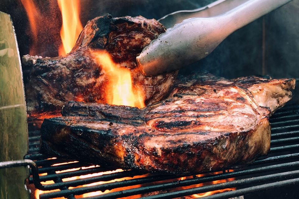 Pedaços de carne bem passada sendo preparada na grelha com fogo alto. No destaque, uma pinça vira um dos pedaços - Metrópoles
