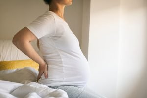Mulher negra grávida sentada na cama enquanto tem as mãos nas costas, indicando dor na região