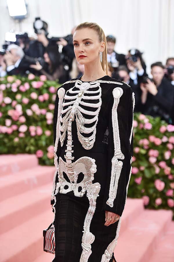 Modelo Carol Trentini na entrada do evento MET Gala. Ela usa um vestido preto com desenhos de um esqueleto branco. Seu cabelo está preso para destacar os brincos de diamante e o batom vermelho.