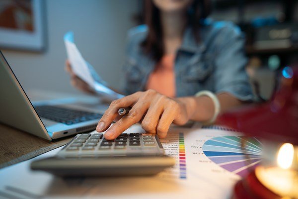 mulher mexe no computador e ao mesmo tempo em que analisa um papel e faz cálculos na calculadora