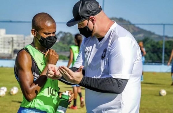 técnico de futebol de boné preto, máscara preta e roupa branca conversa com jogador de colete verde e máscara preta em um treino