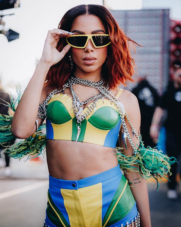 A cantora Anitta, uma mulher morena de cabelos ondulados ruivos, durante o show do festival Coachella. Ela usa óculos escuros com armação amarela e um look composto por top + short, tudo nas cores verde, amarelo e azul, em referência à bandeira do Brasil