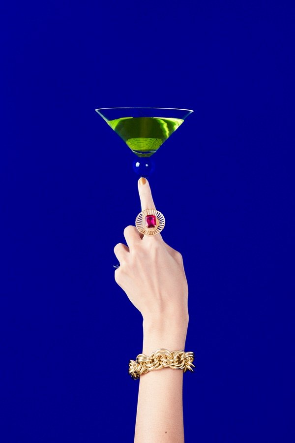 Mão branca segurando uma taça com uma bebida na cor verde. A pessoa usa um anel dourado com pedra rosa e uma pulseira de ouro. O fundo da foto é liso e azul escuro