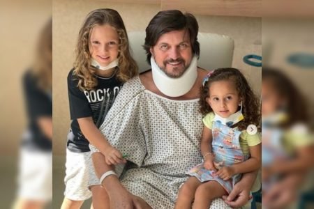 Homem sentado com roupa de hospital e pescoço enfaixado e duas crianças ao lado
