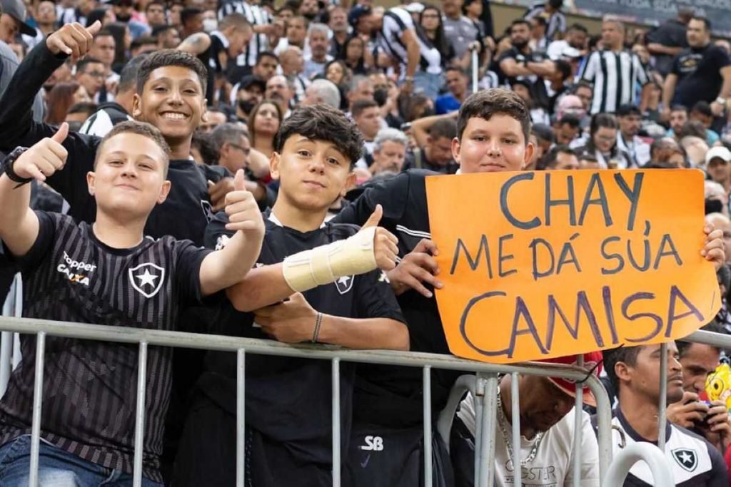Torcedores atrás de grade com roupas pretas. Menino segura cartaz laranja no canto da imagem