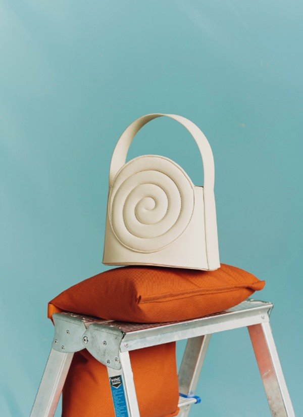 Bolsa branca com um detalhe em aspiral, em um tom de off white. A peça está em cima de uma almoçada laranja, que por sua vez está em um degrau de uma escada de construção. O fundo da foto é azul.