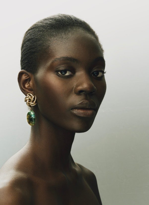 Mulher negra com cabelo amarrado em coque baixo e maquiagem leve. Ela usa um brinco grande, com detalhes dourados, e uma pedra verde poderosa.