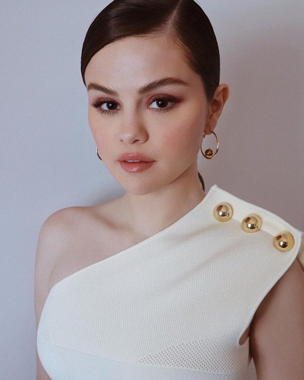 Singer Selena Gomez with her hair tied in a bun. She wears gold hoop earrings and a white dress.