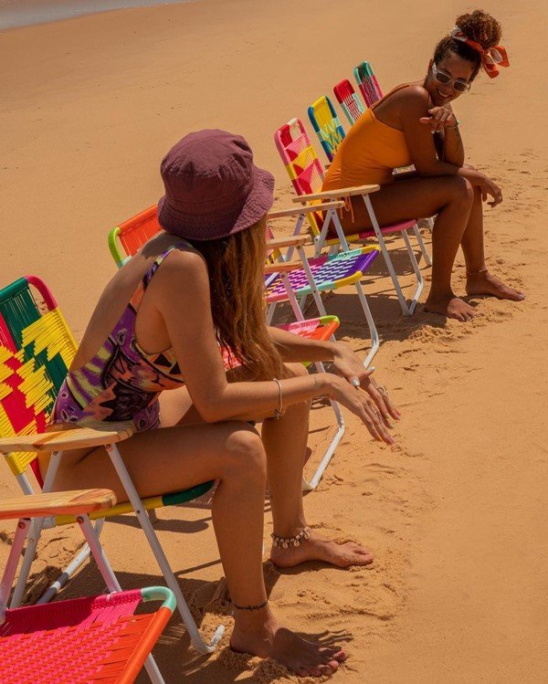 Mulher branca com cabelo liso e louro e mulher negra com cabelo cacheado amarrado em coque conversando na praia. Elas estão sentadas em cadeiras de praia e usam maiôs coloridos