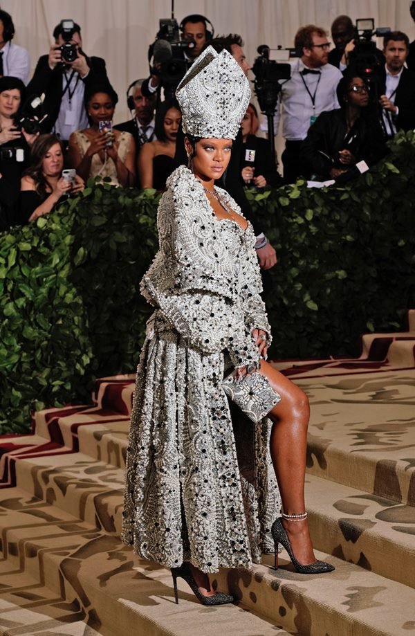 Cantora Rihanna com vestes que lembram as roupas de um papa. As peças são brancas com bordados prateados. A roupa é da marca Maison Margiela. Ela está no tapete vermelho do evento Met Gala.