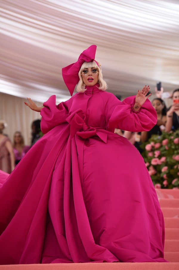 Cantora Lady Gaga com peruca de cabelo louro, quase branco, curto. Ela usa um vestido rosa gigante, com laços e mangas bufantes. Na cabeça, usa um laço rosa, também grande. Ela está na entrada do Met Gala.