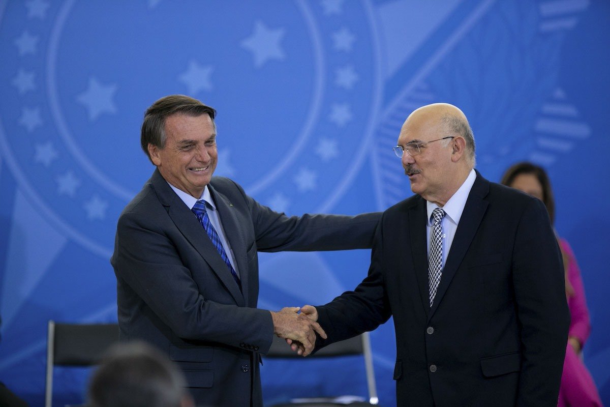 Jair Bolsonaro e Milton Ribeiro se cumprimentam com um aperto de mãos. Os dois usam terno e sorriem