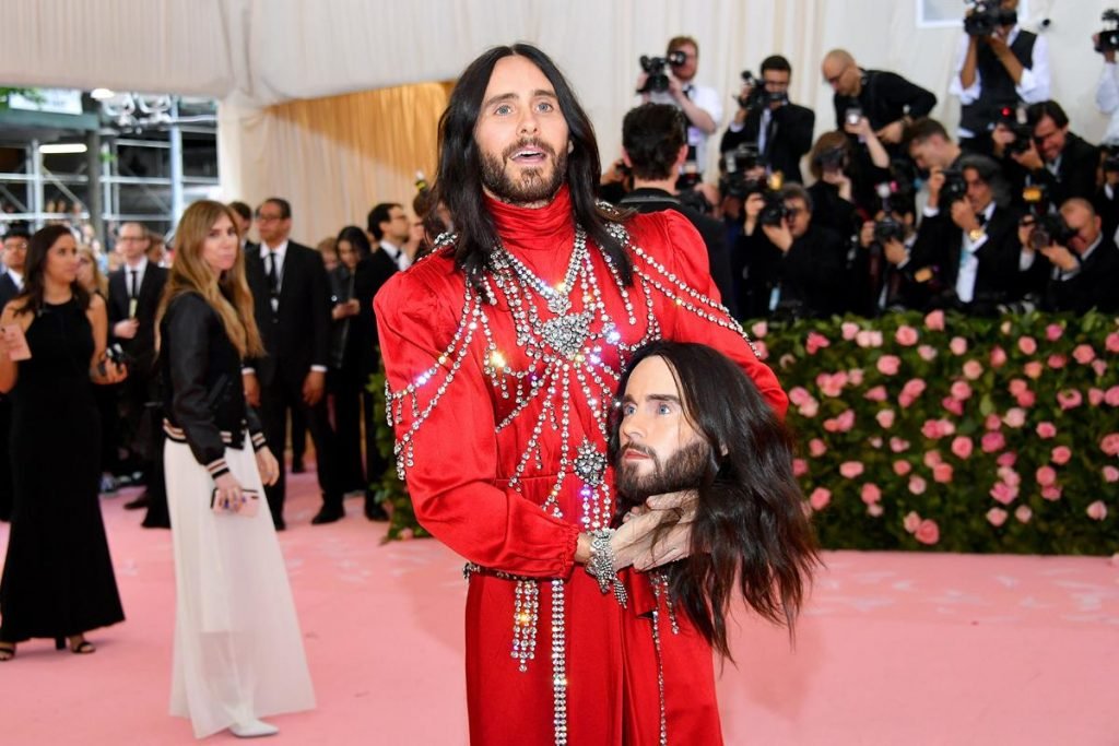 Cantor e ator Jared Leto com vestido azul e, por cima, adornando, correntes prateadas e com brilho. Ele segura, com as duas mãos, uma cabeça cenográfica que imita a sua própria face. A roupa é da marca Gucci e o cantor está no tapete vermelho do baile do Met Gala.