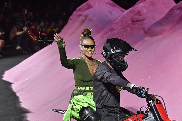 Cantora Rihanna andando em cima de uma moto, com um motoqueiro dirigindo, no desfile da marca Puma. Ela usa uma blusa verde de manga cumprida, um shorts e óculos escuros