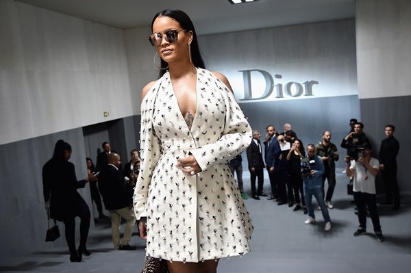 Cantora Rihanna de vestido branco com glitter e óculos escuros espelhados. Ela está em um desfile da marca Dior.