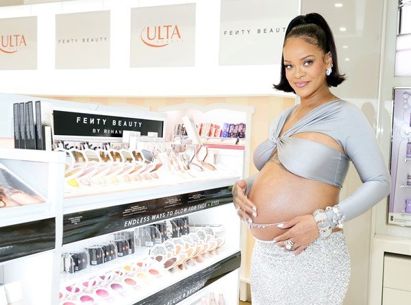 Cantora Rihanna encostada em um balcão de maquiagens da sua marca, Fenty Beauty, em farmácia nos Estados Unidos. Ela está grávida e usa o cabelo amarrado em um rabo de cavalo. Veste um top prata, curto, que deixa a barriga à mostra, e uma saia brilhante, também prateada.