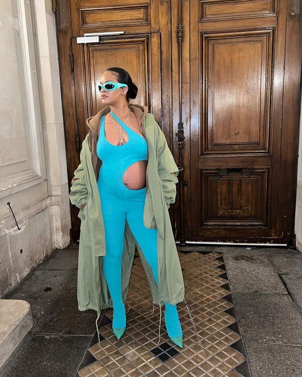 Cantora Rihanna posando para foto na entrada de uma casa. Ela está grávida e usa um macacão azul piscina, colado ao corpo, e com recortes na barriga e no top. Usa, ainda, um óculos escuros com armação na mesma cor da roupa.