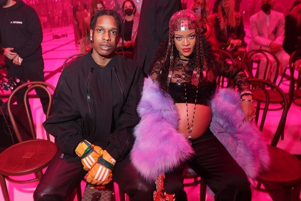 ASAP Rocky e Rihanna sentados no desfile da Gucci. Ele usa camiseta, casaco e calça, tudo preto, e luvas laranjas. Ela usa um top preto, deixando a barriga à mostra, com um casaco de pelos lilás e uma calça preta.