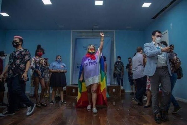 pessoas reunidas em uma sala. no meio, uma pessoa levanta os braços com o punha cerrado. ela leva uma bandeira LGBT nas costas