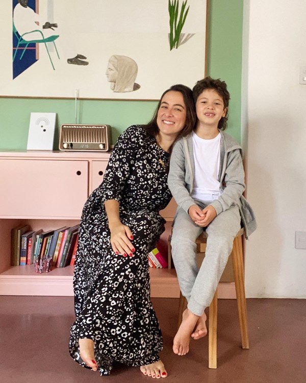 Mulher branca com cabelo castanho, sentada em um banco, ao lado do filho, um menino branco de cabelo escuro com aproximadamente 5 anos