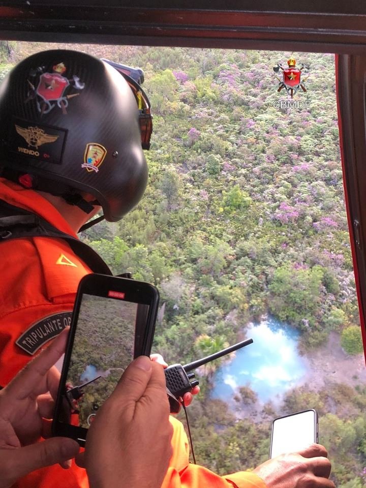 Homem com camisa laranja e capacete preto de costas, em aeronave sobrevoando floresta