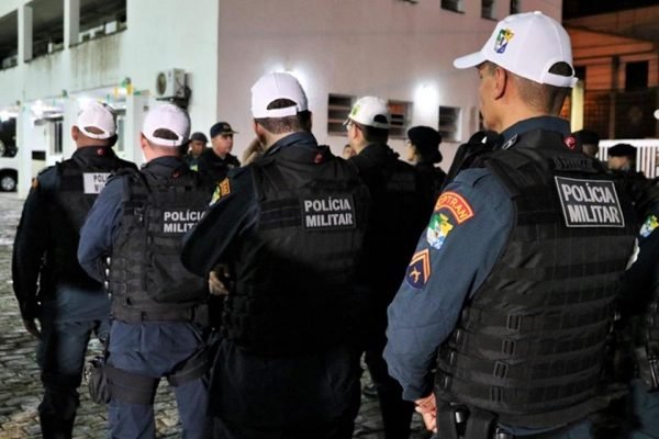 Policiais militares de Sergipe enfileirados e de costas em área urbana, de noite. Todos usam uniforme, colete à prova de balas e bonés brancos - Metrópoles
