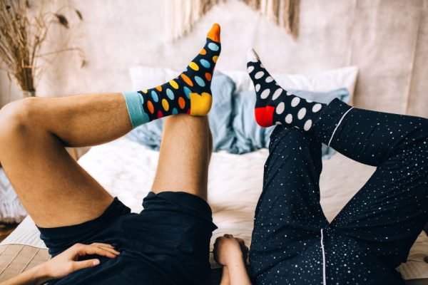 Duas pessoas deitadas numa cama, de mãos dadas, usam meias coloridas e divertidas de bolinhas - Metrópoles
