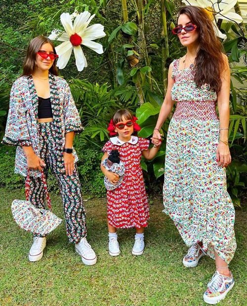 Mulher branca com cabelo liso, castanho, posando para foto no jardim com suas duas filhas. Todas usam roupas estampadas de flores